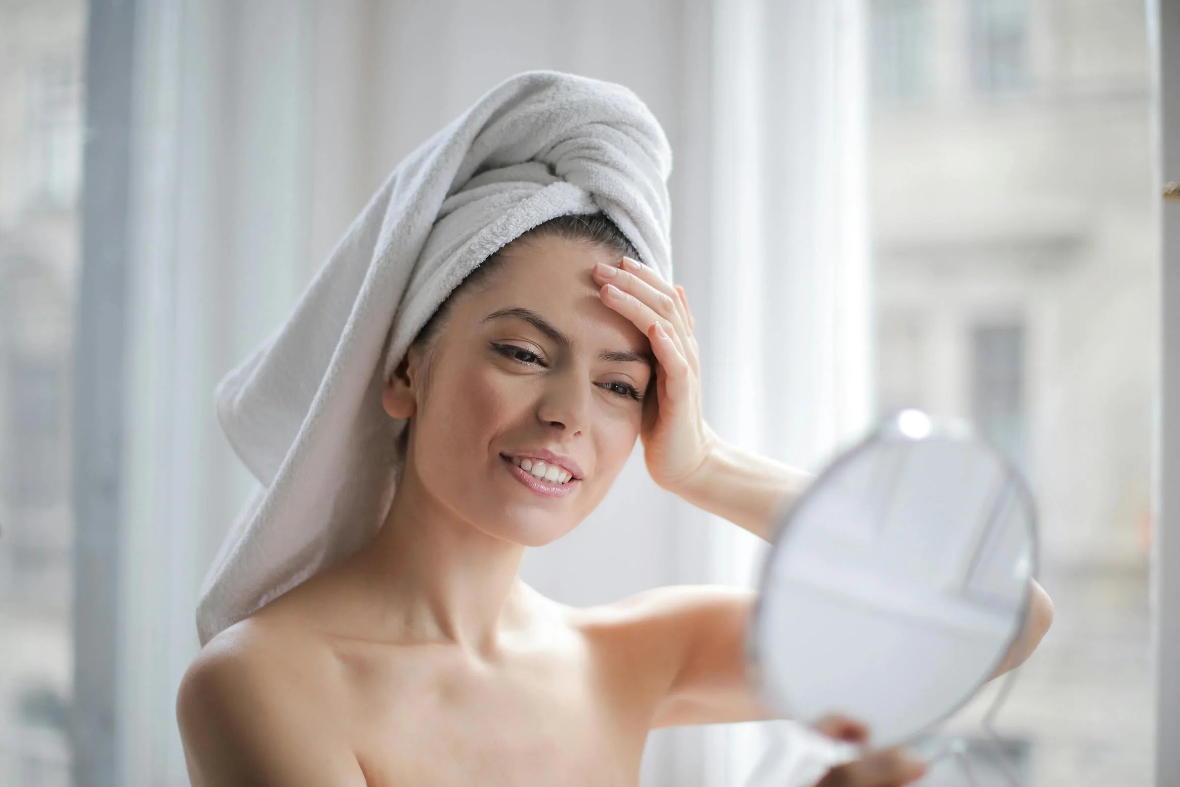 Woman with a towel on her head smiling while looking in a mirror after skincare routine
