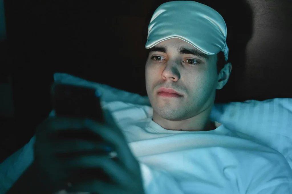 Man in bed wearing a sleep mask looking at phone screen late at night, exposed to blue light