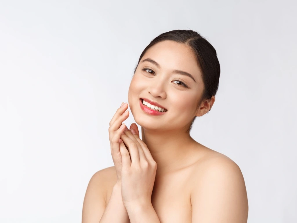 Young Asian woman with glowing skin and natural makeup, smiling while touching her cheek against a white background.