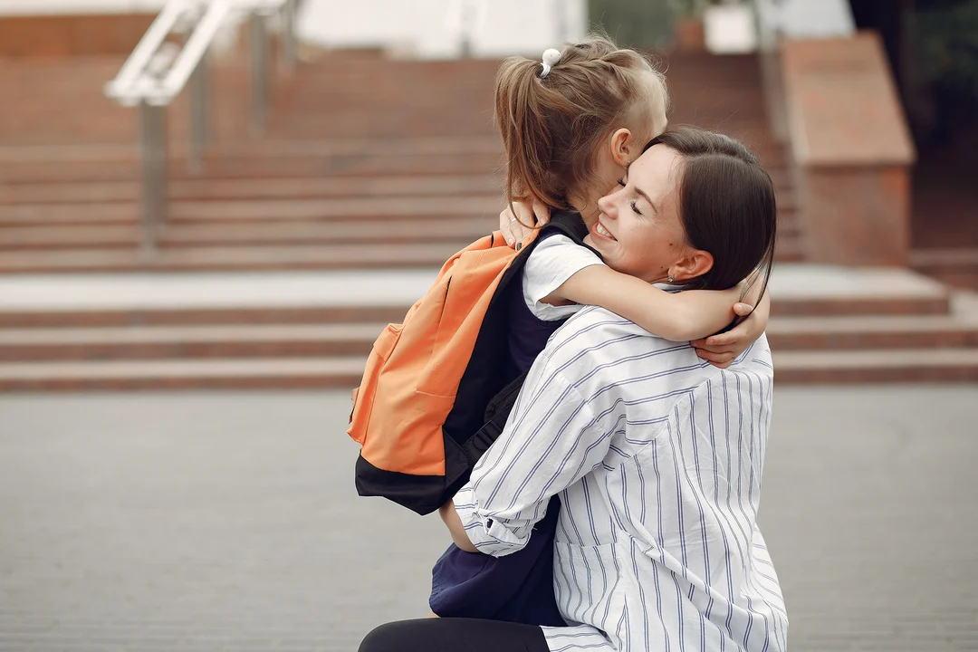 Mother warmly hugging her young daughter with a school backpack, smiling before the child heads to school.